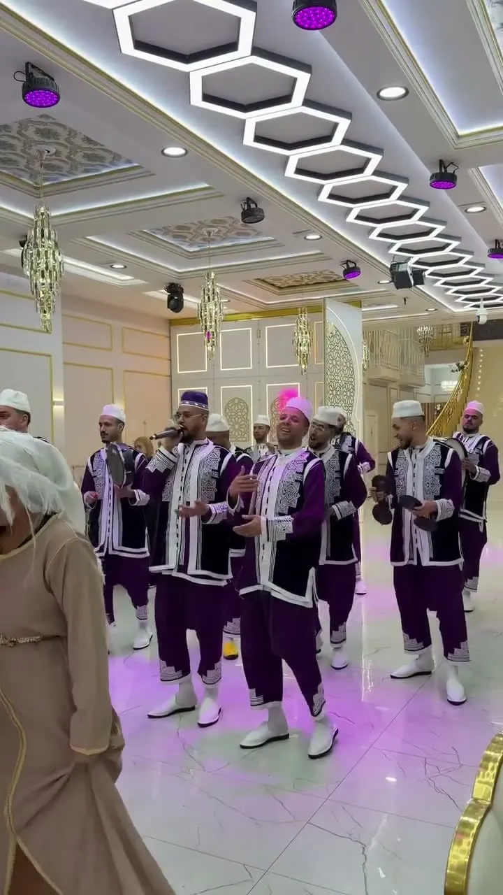 Grupo de músicos con jabadores morados y blancos cantando y tocando panderos en un salón con lámparas hexagonales